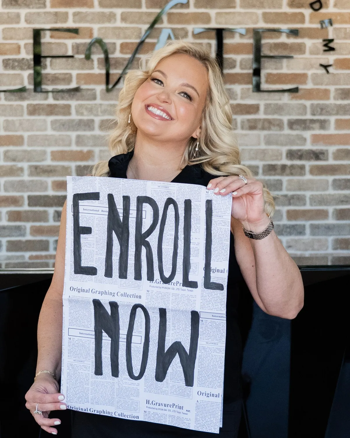 Kendra Waterhouse, director of Elevate Academy, holding an Enroll Now sign in front of the academy's brick feature wall